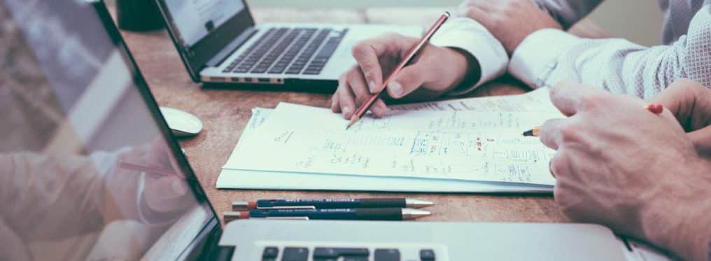 Employees writing on piece of paper that is positioned between two macbook computers.
