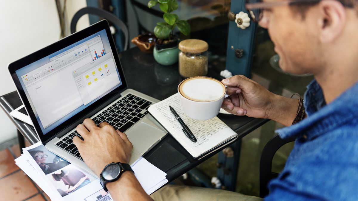 A man holding coffee is seated outside at his desk in front of his computer doing work
